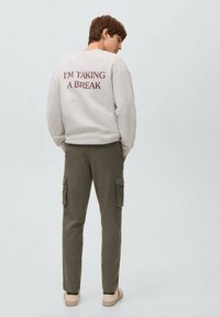 Grå sweatshirt med "JAG TAR EN PAUS" tryckt i mörkröd färg på ryggen, tillsammans med olivgrön cargobyxor och beige slipskor.