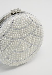 Okrągła torebka na kopertę wykonana z białej tkaniny, ozdobiona perełkowymi koralikami i srebrnymi akcentami; ma geometryczny wzór o gładkiej fakturze.
