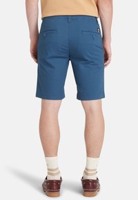 Bermuda in cotone blu, lunghezza al ginocchio, con due tasche posteriori dotate di chiusura a bottone. Indossati con calze beige e scarpe da barca marrone scuro.