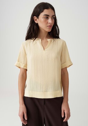Femme aux longs cheveux foncés portant un chemisier texturé jaune clair à manches courtes et un pantalon foncé, regardant vers la gauche sur un fond uni.