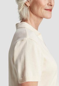 Femme âgée avec des cheveux blancs courts, portant un haut en tricot crème à manches courtes avec un col côtelé et des boucles d'oreilles en or.