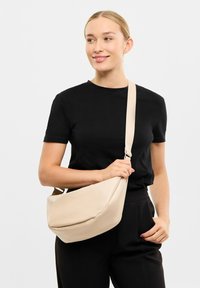 Bežová crossbody taška s křivkovým designem, hladkým povrchem a nastavitelným popruhem, kterou drží osoba v černém tričku na jednobarevném pozadí.