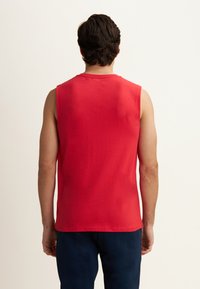 Hombre con cabello oscuro que lleva una camiseta sin mangas roja y pantalones oscuros, de pie de espaldas frente a un fondo claro y sencillo.