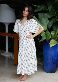 Femme en robe blanche à pois se tenant pieds nus à l'intérieur, posant les mains sur les hanches à côté d'un grand pot bleu avec de grandes feuilles vertes.