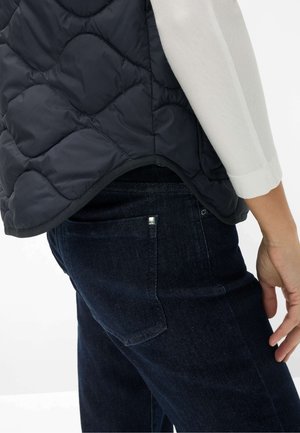 Person wearing dark quilted vest over a white long-sleeve shirt and dark blue denim jeans, showing backside and right hand.