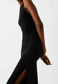 Černé šaty bez rukávů s bočním rozparkem a těsně přiléhajícím střihem. Vyrobené z hladké tkaniny s mírným leskem.