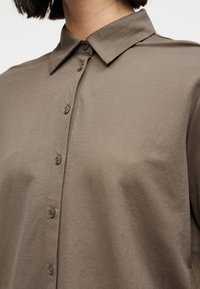 Καφέ πουκάμισο με κουμπιά και κλασικό γιακά που φοριέται από ένα άτομο, αναδεικνύοντας την υφή του υφάσματος και τις λεπτομέρειες των κουμπιών.