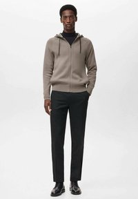 Lichte beige zip-up hoodie van zachte stof met een gladde textuur, voorzien van een trekkoordcapuchon. Draagt met zwarte op maat gemaakte broeken en zwarte schoenen.