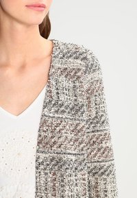 Strickjacke mit Textur, die ein Muster aus grauen, schwarzen und braunen geometrischen Formen zeigt. Offenes Design mit einer weichen, gewebten Textur.