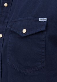 Camicia blu marino con design a bottoni, dotata di una tasca sul petto, bottoni dorati e dettagli di cucitura a contrasto. Etichetta visibile sulla tasca.