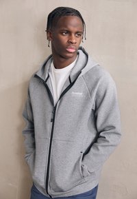 Junger Mann mit geflochtenem Haar, trägt einen grauen Kapuzenpullover mit Reißverschluss und ein weißes Hemd, steht vor einer schlichten beigen Wand, Hände in den Taschen.