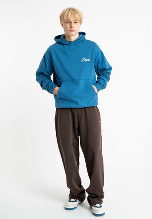 Niebieska bluza z kapturem z napisem "Japan", przednią kieszenią i sznurkiem do kaptura. Brązowe spodnie dresowe. Model nosi szaro-czarne sneakersy z niebieskimi akcentami.