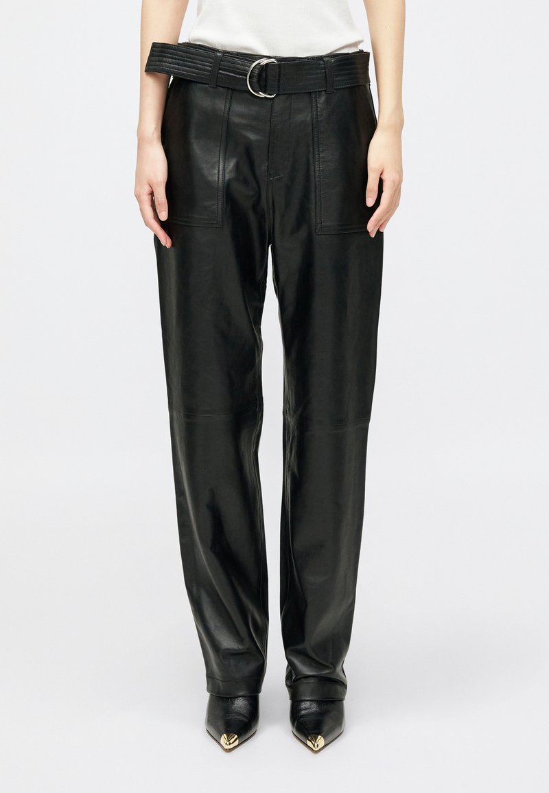 Μαύρα δερμάτινα παντελόνια ψηλής μέσης με μπροστινές τσέπες, ευρύς ζώνη με ασημένιο κούμπωμα, συνδυασμένα με μυτερές μαύρες μπότες με χρυσές άκρες.