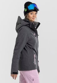 Grijze skijack met roze ritsen en accenten, voorzien van een capuchon en verstelbare manchetten, gecombineerd met roze skibroeken en een zwarte beanie.