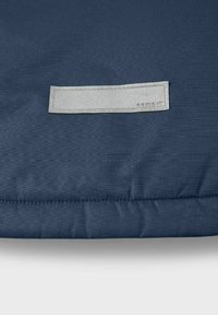 Tmavě modrý polštář s hladkým povrchem a prošitými okraji. Obsahuje světle šedou nálepku s vyraženým textem pro branding.