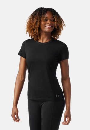 Donna sorridente con capelli ricci che indossa una maglia nera a maniche corte e leggings neri, in piedi davanti a uno sfondo bianco semplice.