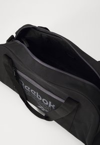 Bolsa de deporte negra con cremallera superior, tela texturizada, bolsillo lateral y logo de Reebok en gris. El interior es amplio con un pequeño bolsillo con cremallera.