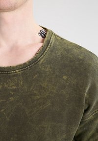 Olivgrön, urtvättad bomullströja med rundad halsringning, slitna fållar och subtil textur. Synlig nacktattoo ovanför kragen.