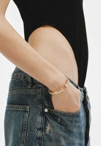 Pulsera de metal dorado con un diseño en espiral, llevada en un brazo desnudo, combinada con unos vaqueros de denim de cintura alta en un tono azul desgastado. Estética simple y moderna.