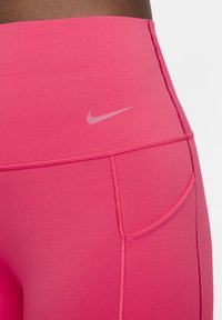 Roosa kõrge vöökohaga retuusid, millel on sile ja elastne kangas, küljetasku ning peen Nike logo sobivas roosas toonis vööl.