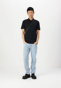 Camisa polo preta, jeans azul claro e sapatos pretos de sola grossa. A camisa tem um colarinho clássico; os jeans têm um corte mais descontraído.