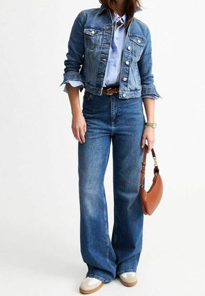 Kobieta ubrana w przyciętą jeansową kurtkę, jasnoniebieską koszulę na guziki, jeansy z wysokim stanem i szerokimi nogawkami, białe trampki, trzymająca brązową torebkę.
