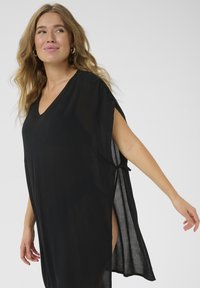 Kaftán negro hecho de una tela ligera y transparente. Presenta un escote en V, mangas cortas y lazos en los laterales con aberturas para facilitar el movimiento.