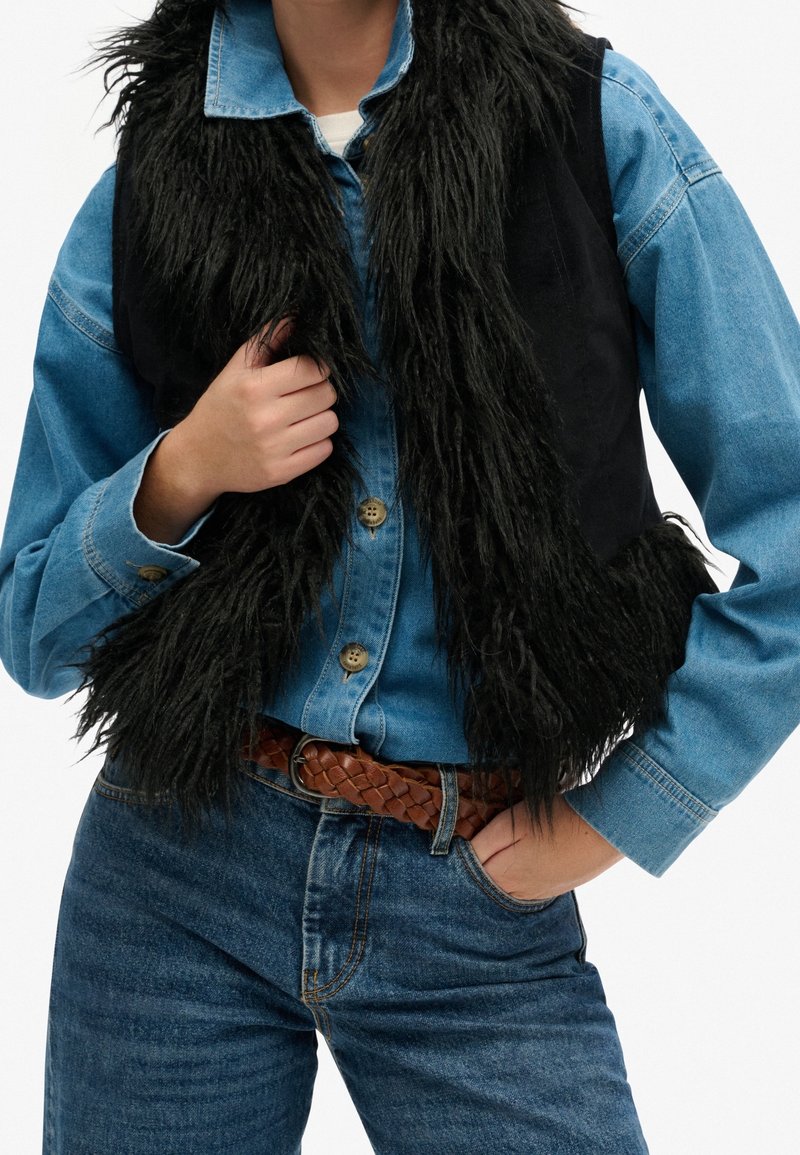 Czarna kamizelka ze sztucznego futra nałożona na niebieską dżinsową koszulę, zestawiona z ciemnoniebieskimi dżinsami i brązowym plecionym paskiem ze skóry.