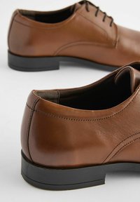 Chaussures habillées en cuir marron avec une texture lisse, un bout arrondi et des semelles en caoutchouc noir. Elles présentent des lacets et un détail cousu sur le côté.