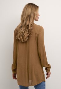 Blusa a maniche lunghe, marrone, trasparente, con tessuto testo, morbido drappeggio e polsini con bottoni; abbinata a jeans azzurri.