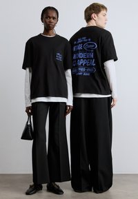 Čierne nadrozmerné tričká s modrými grafickými potlačami, nosené cez biele tričká s dlhým rukávom. Čierne nohavice s širokými nohavicami a čierna kabelka dotvárajú vzhľad.