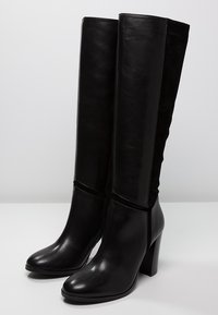 Botas negras hasta la rodilla que combinan cuero suave y tela texturizada, con una punta redonda y un tacón robusto.