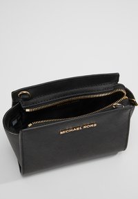 Černá kožená kabelka se zlatými detaily, zipovým uzávěrem a logem "Michael Kors" na přední straně. Interiér má členěný design.