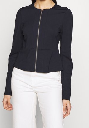 Kobieta ubrana w dopasowaną czarną kurtkę z długim rękawem i zamkiem błyskawicznym, z detalem peplum, w połączeniu z wysokim stanem białych spodni.