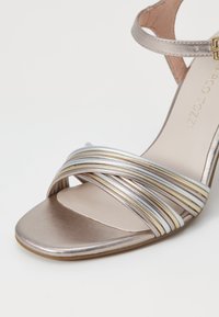 Sandal de tacón metálico con punta cuadrada, que presenta tiras entrelazadas en plata, oro y blanco, y una elegante correa para el tobillo. Textura suave.
