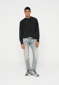 Fiatal férfi áll fekete pulóverben, világos farmernadrágban, fehér-fekete sportcipőben, és jellegzetes övcsattal, fehér háttér előtt.