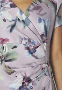 Blommig klänning med en lila bakgrund, prydd med stora pastellblommor i grönt, rosa och blått, insamlad design vid midjan, mjuk tygtextur.