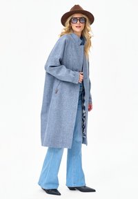 Kobieta ubrana w niebieski wełniany płaszcz, rozkloszowane jasnoniebieskie dżinsy, czarne spiczaste buty, duże okulary przeciwsłoneczne i brązowy kapelusz z szerokim rondem, stojąca na białym tle.