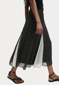 Jupe maxi à pois noir et blanc avec une texture transparente, dotée de fentes sur les côtés et d'un panneau uni, assortie à des sandales plates noires.