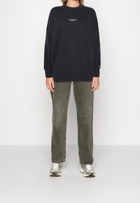 Persona che indossa un felpa nera Abercrombie and Fitch, jeans dritti grigi sbiaditi e sneakers bianco-grigie, in piedi contro uno sfondo bianco.