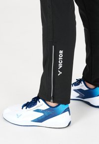 Svarta sportbyxor med siddragkedja, i kombination med vita och blåa träningssneakers med en texturerad design och logotypdetaljer.