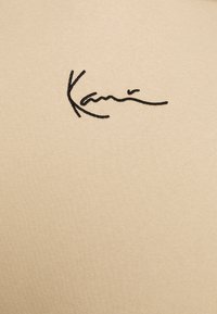 Tessuto di cotone beige con un design di firma ricamato in nero, texture liscia e forma aderente.