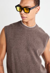 Haut en tricot sans manches marron avec un col rond, tissu texturé, associé à des lunettes de soleil rectangulaires noires avec des verres jaunes.