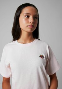 T-shirt en coton rose clair avec un col rond, présentant un petit graphisme brodé d'une fraise et du texte sur le côté gauche de la poitrine.