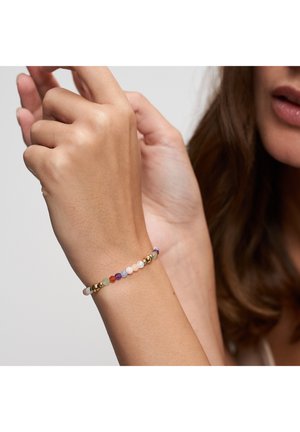 Woman adjusting a beaded bracelet with multicolored and gold round beads on her wrist against a neutral background.
