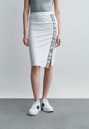 Mujer con una falda blanca hasta la rodilla con texto negro "Emporio Armani" a lo largo de un lado y zapatillas blancas con detalles negros.