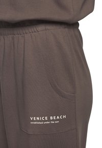 Brown jogger pants featuring an elastic waistband, side pockets, and printed text "VENICE BEACH established under the sun" in white.