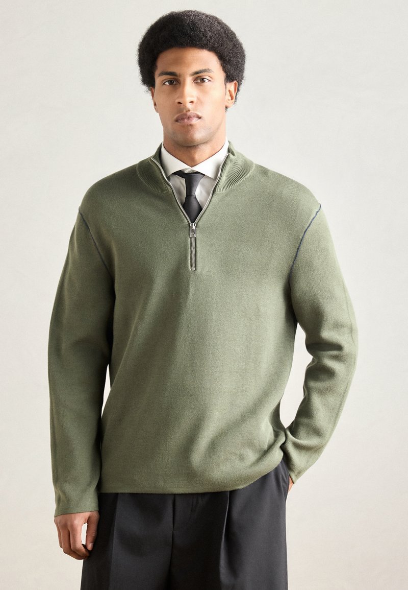 Πλεκτό πουλόβερ σε ελιά πράσινο με γιακά μισού φερμουάρ, ριμπ μανσέτες και άνετη εφαρμογή, σε συνδυασμό με μαύρο παντελόνι ραμμένο στα μέτρα.