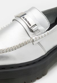 Mocassin en cuir argenté avec semelle épaisse noire à rainures et boucle en métal gravée du logo "Dr. Martens" sur le dessus.