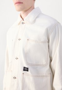 Light beige jacket made of denim fabric, featuring a pointed collar, button closures, and contrasting stitching. Left chest pocket with label.
